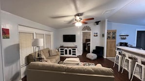 Ein Wohnzimmer mit Sofa, Deckenventilator, Fernseher und einer Küchenzeile mit Barhocker.