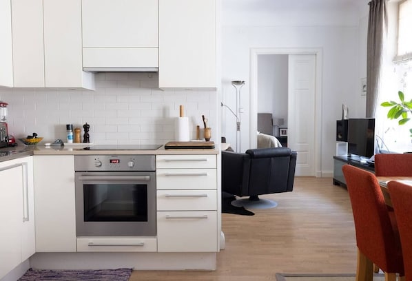 A modern kitchen with white cabinetry, a built-in oven, and a dining area with a black chair and red table.
