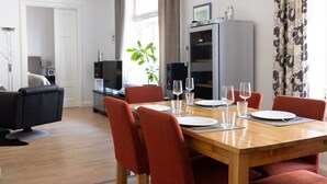Uma sala de jantar com uma mesa de madeira posta para quatro pessoas, cercada por cadeiras vermelhas. Há uma televisão e uma planta ao fundo.