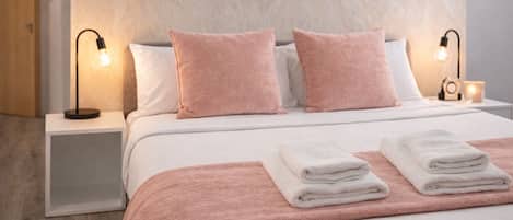 A neatly made bed with pink and white bedding, two bedside lamps, and a stack of folded towels.