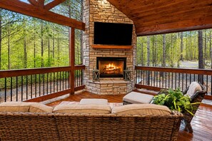 A stone fireplace with a fire, a flat-screen TV above it, wicker furniture, and a view of a forest.