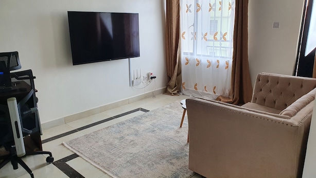 Una sala de estar con una televisión de pantalla plana montada en la pared, un sofá, una mesita, una alfombra y una ventana con cortinas.