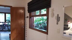 A hotel room with a wooden floor, a window with a view of greenery, a cactus decoration, and a bed with a nightstand.
