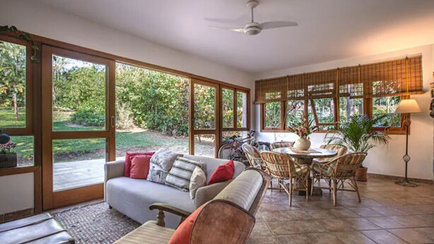 A spacious living area with a sofa, chairs, and a dining table. Large windows provide views of a garden.