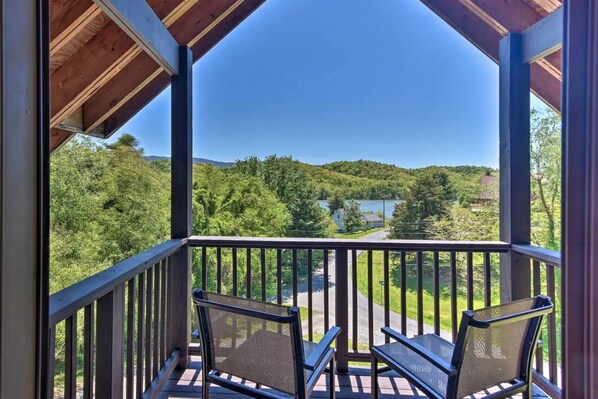 Un espace extérieur abrité doté d’une terrasse en bois, de deux chaises et offrant une vue sur un lac et des collines boisées.