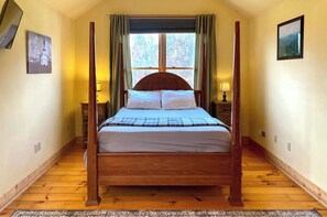 Un lit à baldaquin avec une structure en bois, une literie blanche et une couverture à carreaux. Il y a deux tables de chevet avec des lampes, une fenêtre avec des rideaux et deux tableaux encadrés accrochés aux murs.