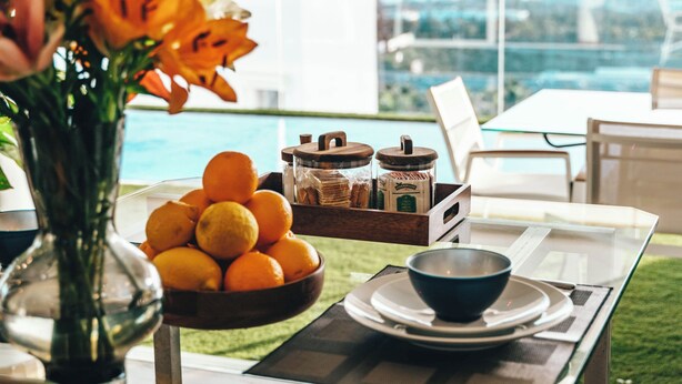 A table with a bowl of oranges, a vase with flowers, and a tray with jars and a box, set on a terrace with a view of the city. - Villa Francia (Costa Adeje)