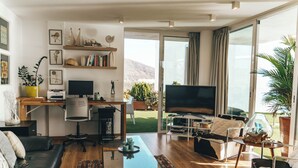 A modern living room with a sofa, coffee table, TV, and a desk with a computer. There are plants, bookshelves, and a view of the outdoors. - Villa Francia (Costa Adeje)