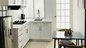 Een kleine keuken met witte kastjes, een gasfornuis, een spoelbak en een eethoek met een tafel en stoelen.