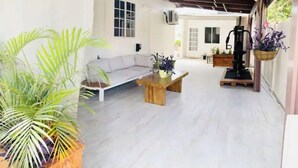 Ruime kamer met een witte bank, een houten salontafel en een potplant. Er is een raam met zonwering, een plafondventilator en een hangplantmand.