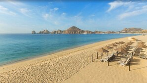 A beach with a row of sun loungers and umbrellas, a clear blue sea, and distant rocky formations. - Beautiful Villa del Arco - Two Bedroom Suite (Cabo San Lucas)