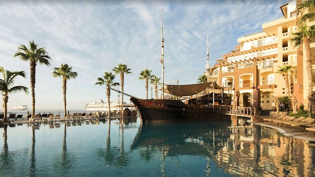 A pool with a boat, palm trees, and a building with balconies. - Beautiful Villa del Arco - Two Bedroom Suite (Cabo San Lucas)