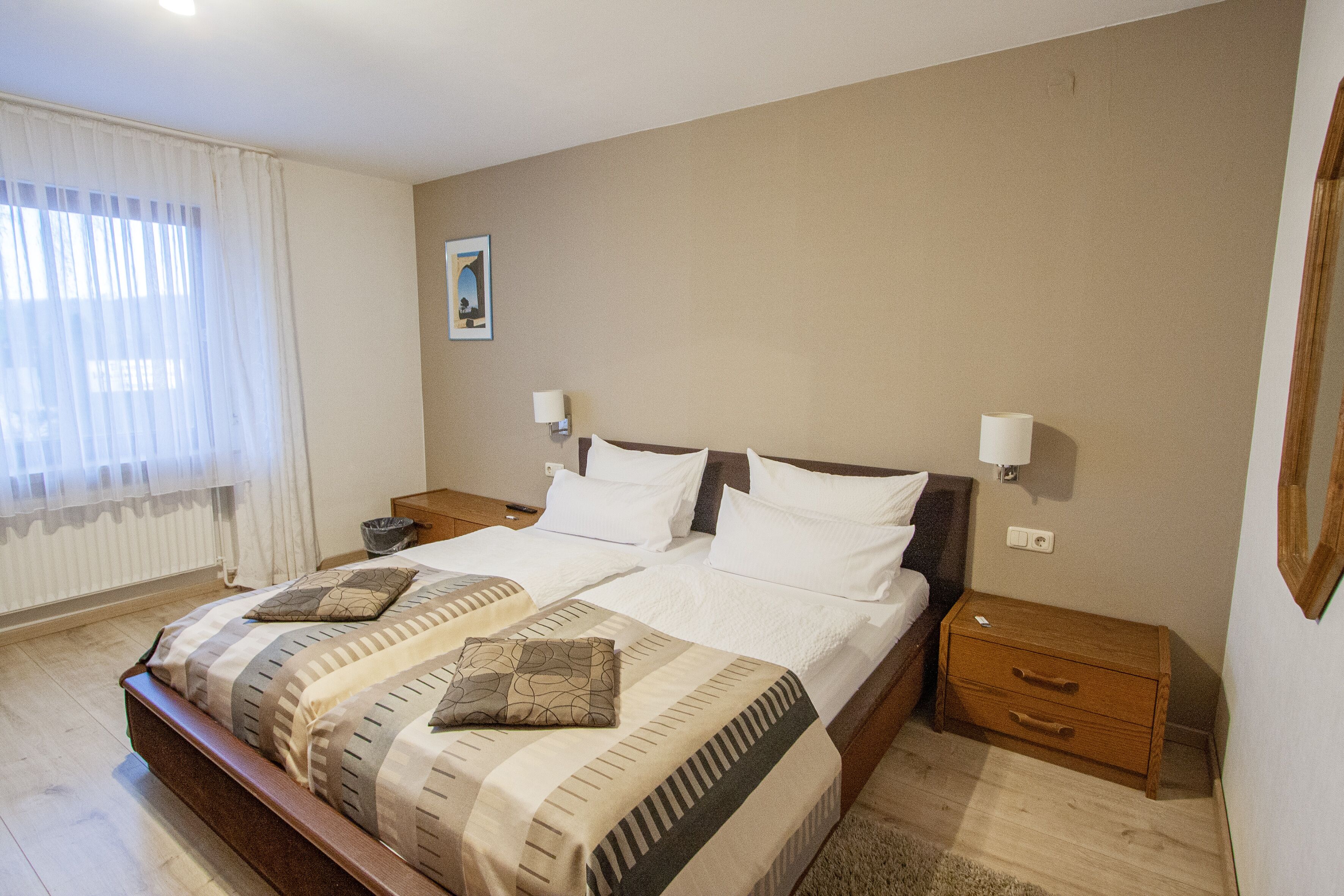A double bed with a striped bedspread, two bedside tables, a nightstand, a wooden dresser, and a window with sheer curtains.