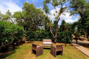 Een terras met rieten meubels en een bank, omringd door weelderige begroeiing en bomen.