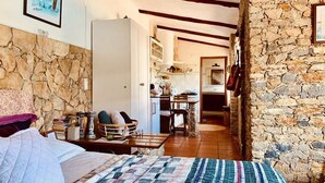 Un dormitorio con cama, mesita de noche y una pared de piedra, además de una cocina al fondo.