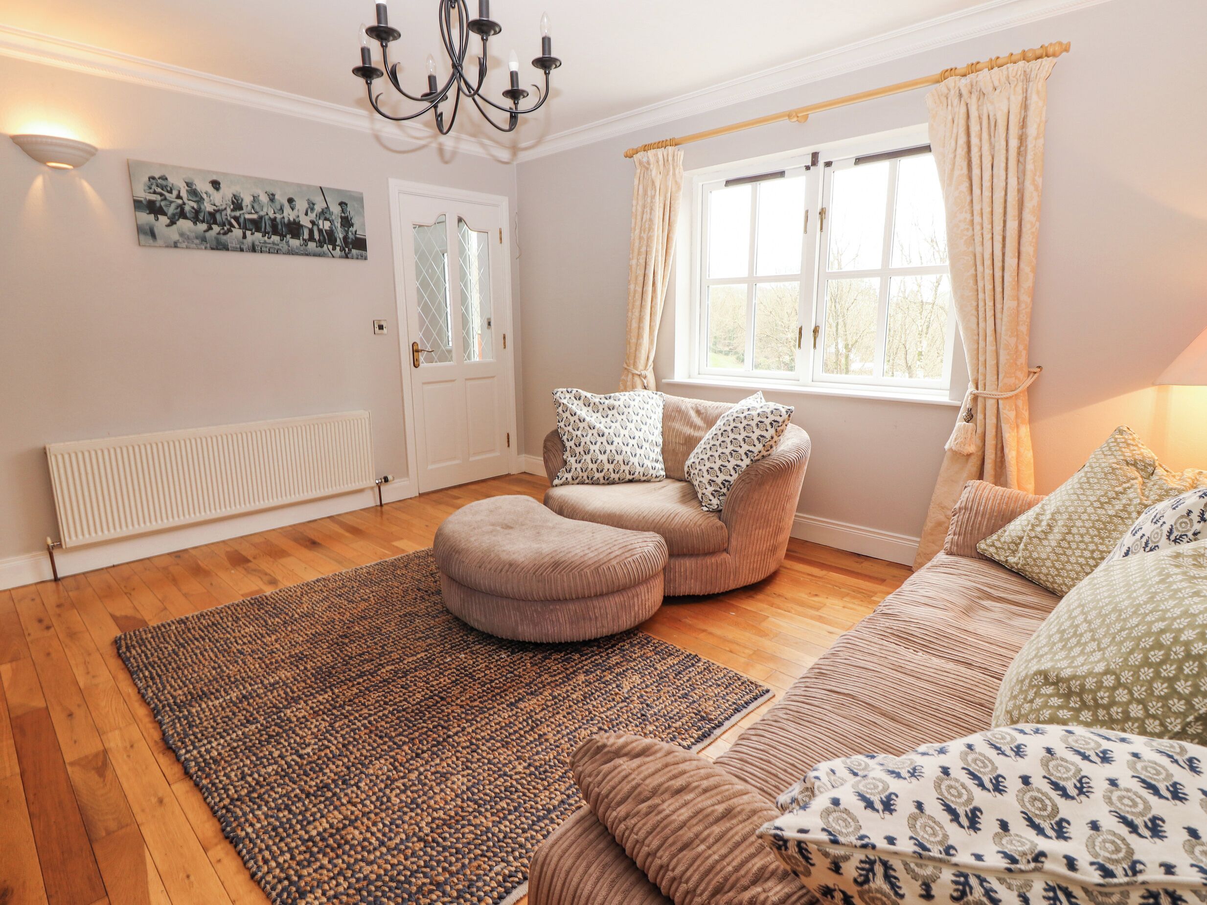 A cozy living room with a beige sofa, a matching ottoman, a patterned rug, a chandelier, and a window with curtains.