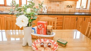 Une cuisine avec des meubles de rangement en bois, un pare-feu en carrelage, un vase blanc rempli de fleurs, un panier contenant du thé et du café, et une assiette de pâtisseries.