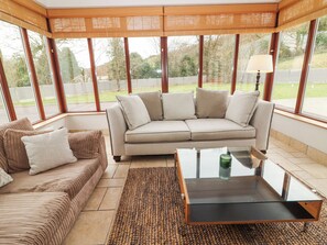 A modern living room with a beige sofa, a patterned sofa, a glass coffee table, and a view of the outdoors through large windows. - Hazelwood (Leap)