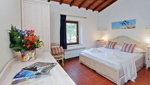 Quarto com cama, mesa de cabeceira, janela com cortinas, radiador, quadro na parede, e uma mesa com um vaso de flores e revistas.