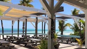 Une terrasse au bord de la plage, avec des tables de pique-nique en bois et un espace abrité offrant une vue sur l’océan.