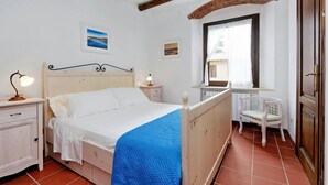 A bedroom with a wooden bed, white walls, a small table with a lamp, a chair, and a window with a view of the outside. - Pimpinnacolo 21 - 1BR P3 8 (Monte Argentario)