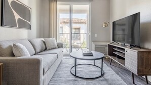 Un soggiorno moderno con un divano grigio, un tavolino rotondo, un televisore a schermo piatto e una porta vetrata scorrevole che dà su un balcone esterno.