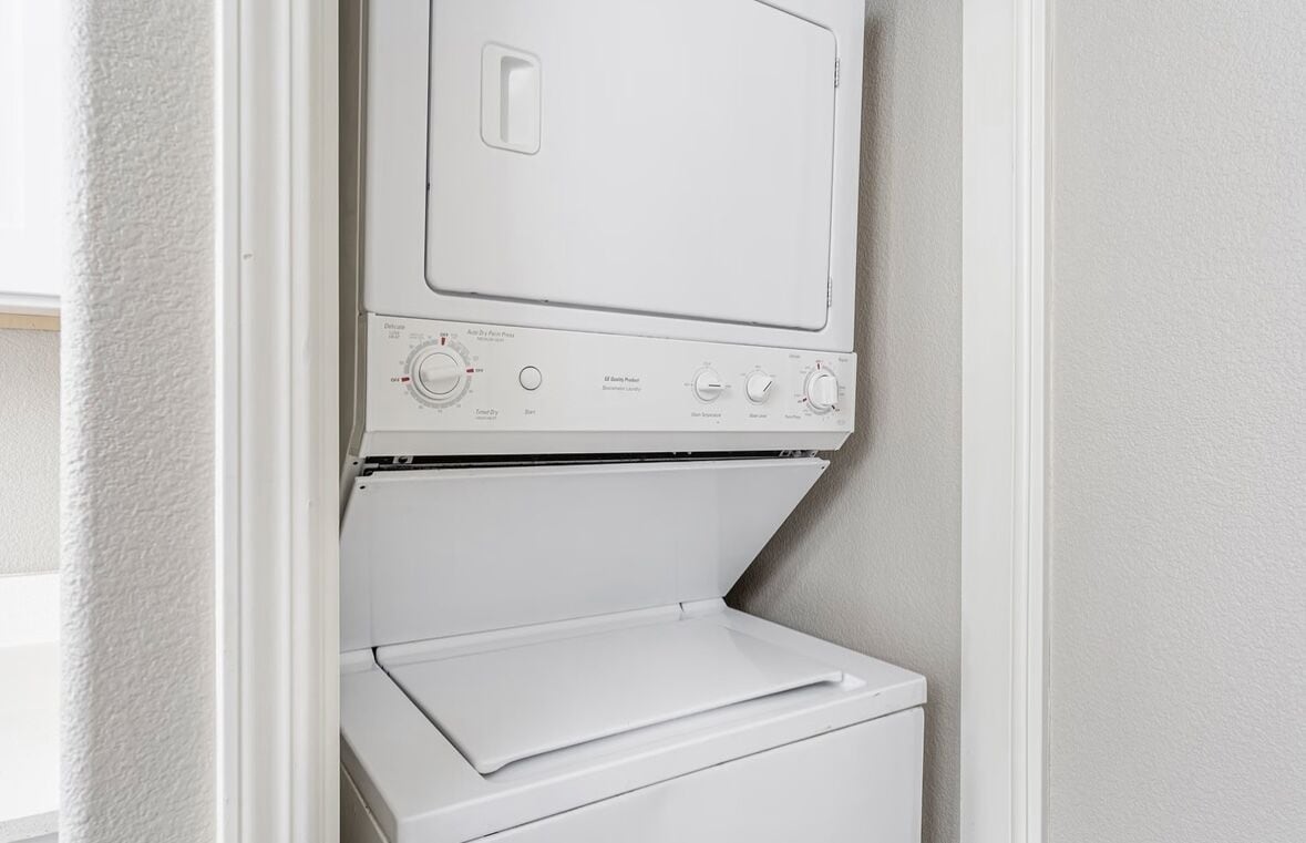 A compact, white laundry room with a front-loading washer and dryer, a small shelf, and a door leading to another room.