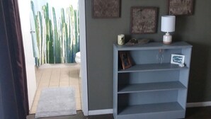 Une chambre d’hôtel avec un lit, un bureau doté d’une lampe, une étagère avec des livres et une tasse, une porte donnant sur un balcon avec vue sur des cactus, et un mur orné d’œuvres d’art encadrées.