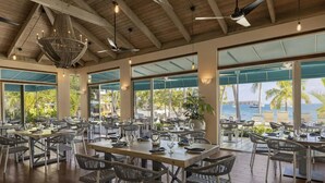 A spacious outdoor dining area with multiple tables and chairs, large windows offering views of the ocean and palm trees, and a rustic wooden ceiling with hanging lights. - Closest to the Sandy Beach/ Direct Ocean front / Full Back up Generator (East End, St Thomas)
