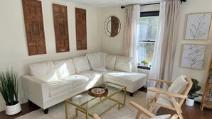 A modern living room with a white sectional sofa, a glass coffee table, and a wooden chair. - Peaceful 2 Bedroom Forest Retreat w/ Sauna! (Ithaca)