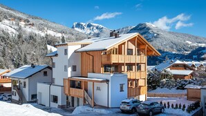 A modern ski resort building with a wooden facade, multiple balconies, and a view of snow-covered mountains.