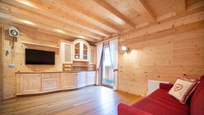 A wooden cabin interior with a kitchen, a TV, a red sofa, and a window with curtains.