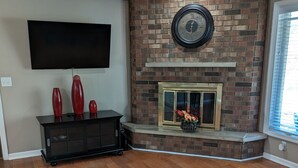 A living room with a brick fireplace, a flat-screen TV mounted on the wall, and a wooden dining table.