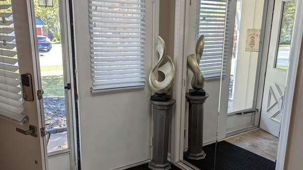 A hallway with a wooden floor, a black doormat, and two abstract sculptures on pedestals.