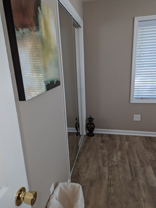 Une pièce dotée d’un plancher en bois, d’une porte-fenêtre coulissante, d’un tableau accroché au mur, d’une fenêtre avec des stores et d’un petit vase décoratif.