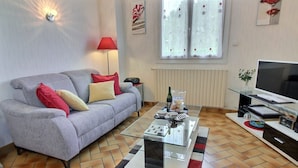 A living room with a grey sofa, a glass coffee table, a TV, and a window with curtains. - Du Gouët - ST Brandan (ST BRANDAN)
