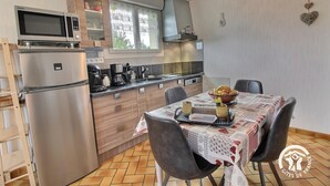 A compact kitchen with a microwave, stove, and refrigerator. There is a dining table with chairs and a fruit bowl. The kitchen has wooden cabinets and a window with lace curtains. - Du Gouët - ST Brandan (ST BRANDAN)