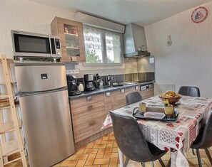 Une cuisine compacte équipée d’un four à micro-ondes, d’une cuisinière et d’un réfrigérateur. On y trouve une table à manger avec des chaises et un panier de fruits. La cuisine est dotée de meubles en bois et d’une fenêtre agrémentée de rideaux en dentelle.