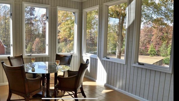 A sunroom with a glass table and four chairs, large windows, and a view of a red barn and trees.