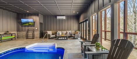 An indoor pool area with a sofa and chairs, a television, and a view of the outdoors through large windows.