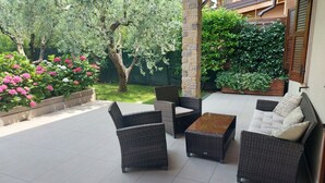 Una terrazza esterna con mobili in vimini, un pilastro in pietra e vista su un giardino con alberi e fiori.