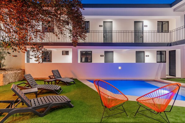 Un edificio de dos pisos con piscina, sillas de descanso y un árbol.