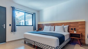 A modern hotel room with a large bed, checkered bed skirt, a wooden headboard, a bedside table with a lamp, and a window with curtains.