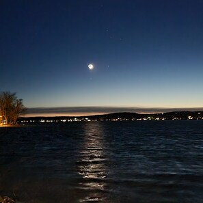 Ein Nachtbild einer Küstenlandschaft mit ruhiger See, einem sichtbaren Mond und fernen Stadtlichtern.