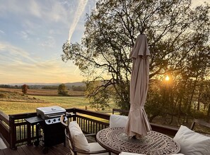 Eine Terrasse mit Grill, Stühlen und Sonnenschirm mit Blick auf eine malerische Landschaft, Bäume und einen Sonnenuntergang.