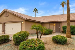 A single-story house with a garage, a front porch, and a landscaped garden with palm trees and shrubs.