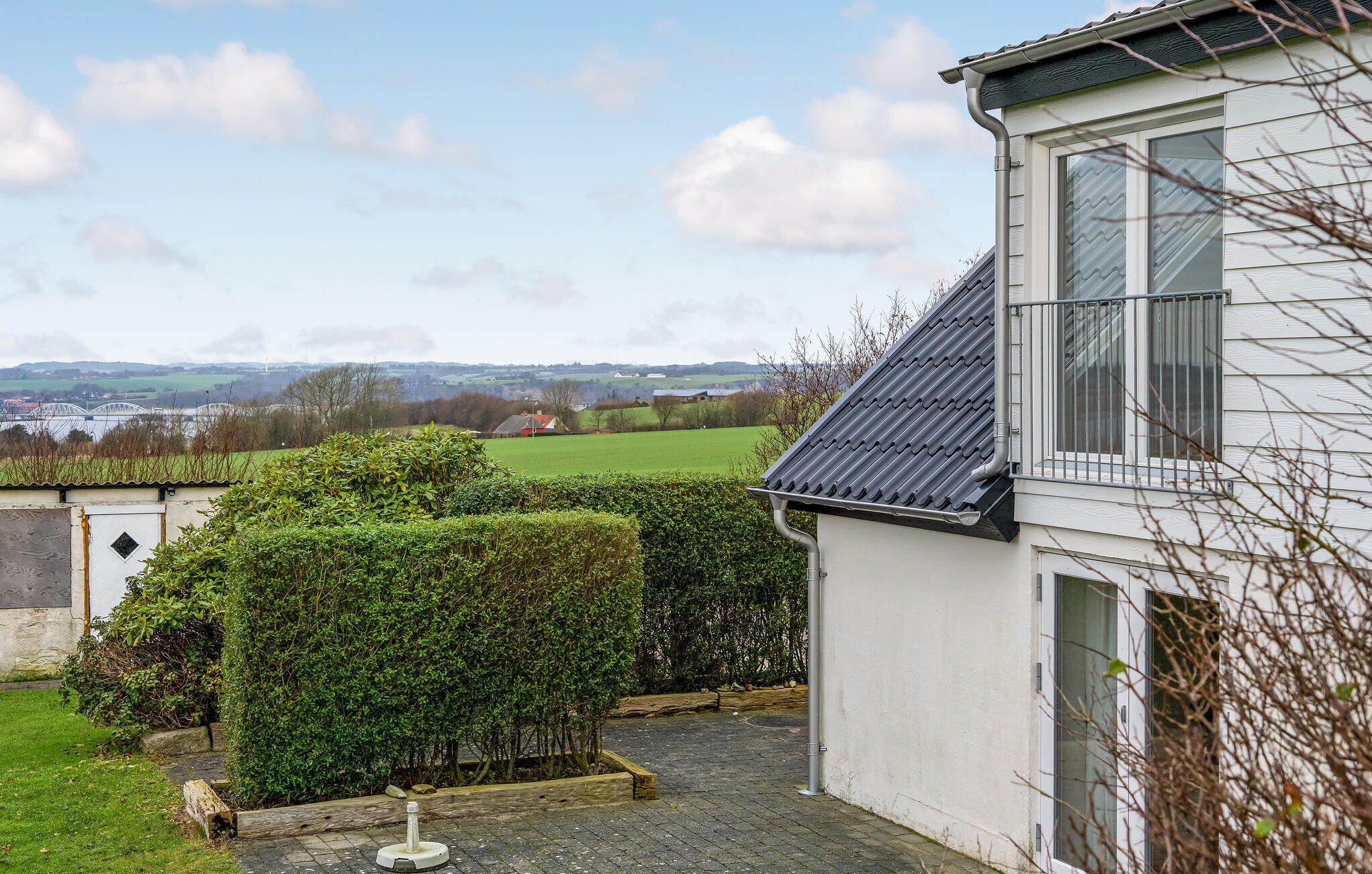 A house with a white exterior, a tiled roof, and a view of a green field and a bridge in the distance.