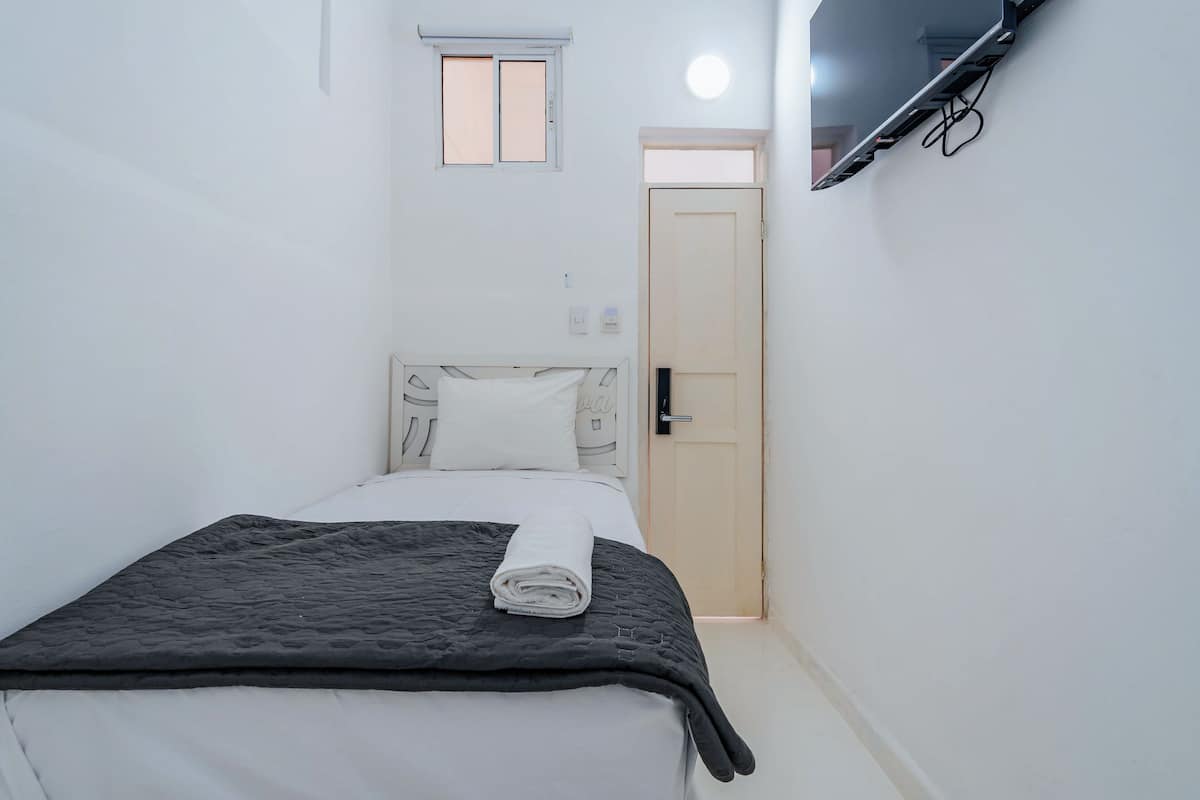Una cama pequeña y bien hecha con una toalla blanca enrollada, una ventana pequeña y una puerta en una habitación minimalista.