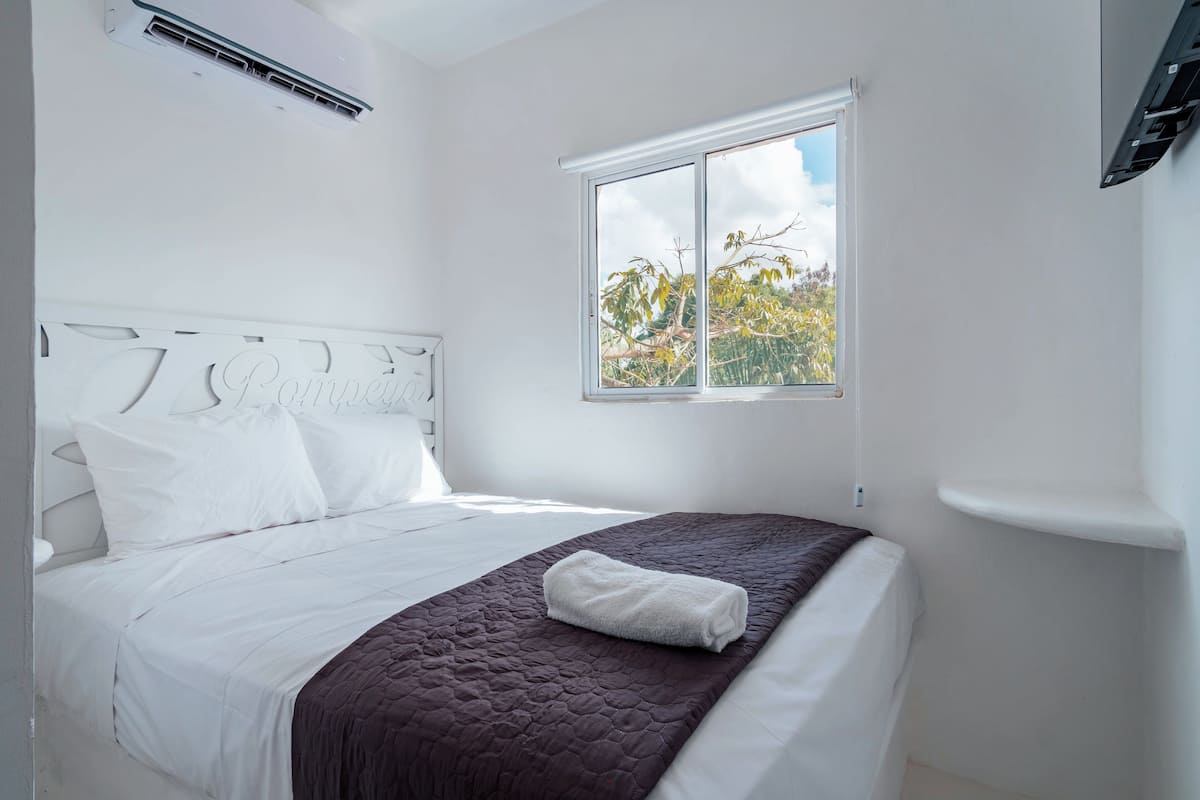 Un dormitorio con cama blanca, ventana con vista a árboles y unidad de aire acondicionado blanca.