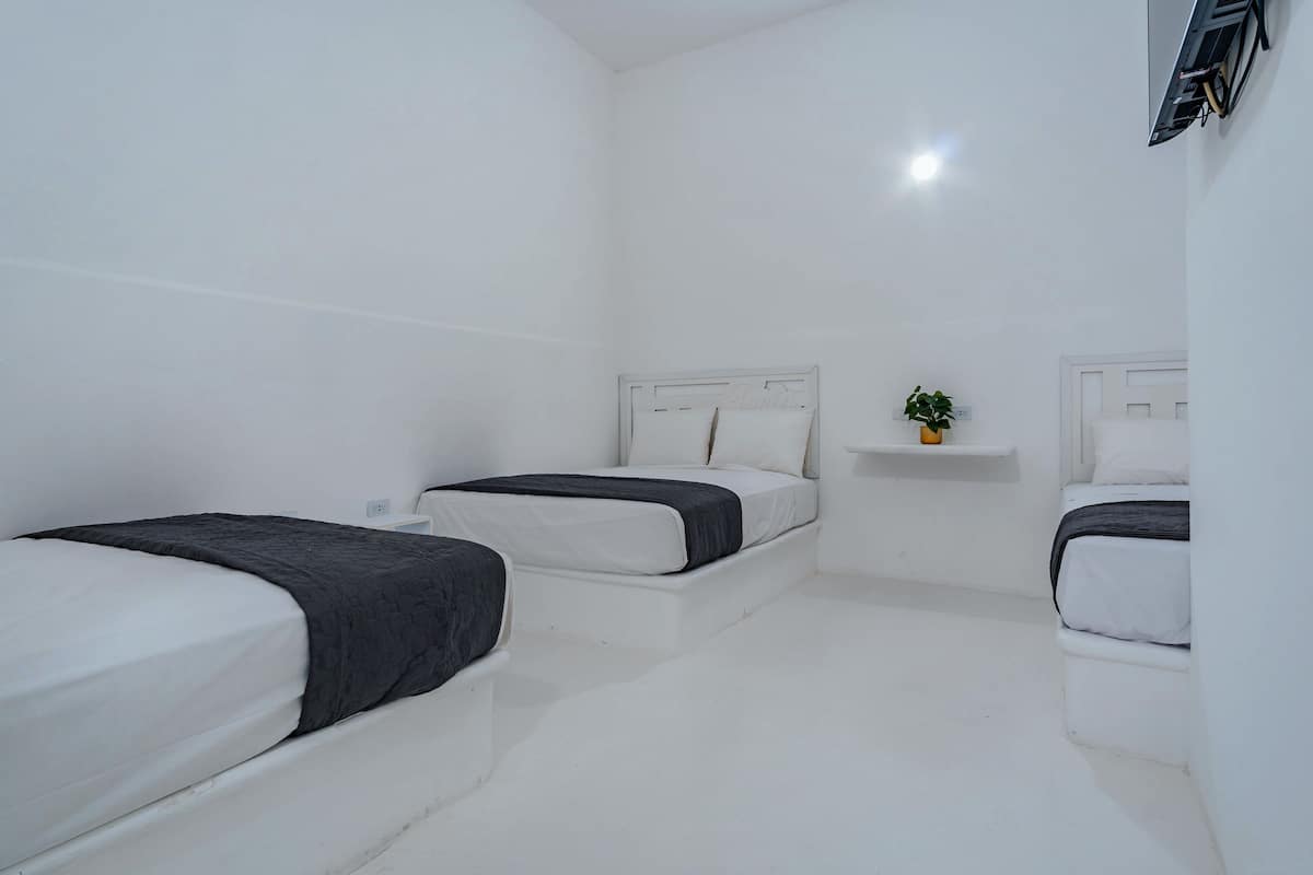 Un dormitorio minimalista con decoración blanca, con dos camas, una mesita de noche con una planta y un aire acondicionado de pared.